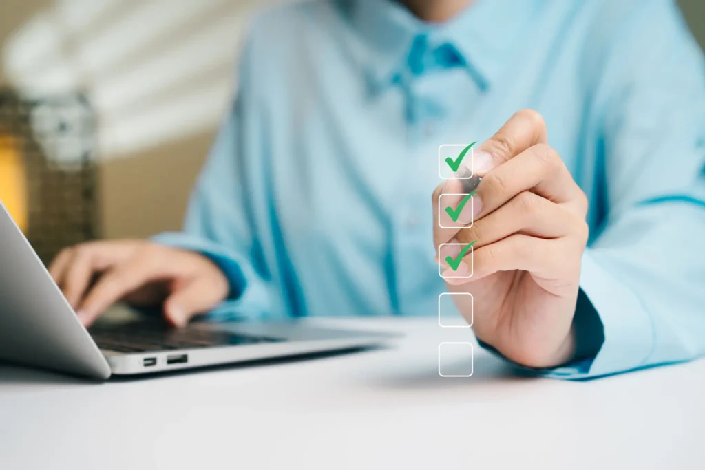 Person checking boxes on a transparent checklist near a laptop.